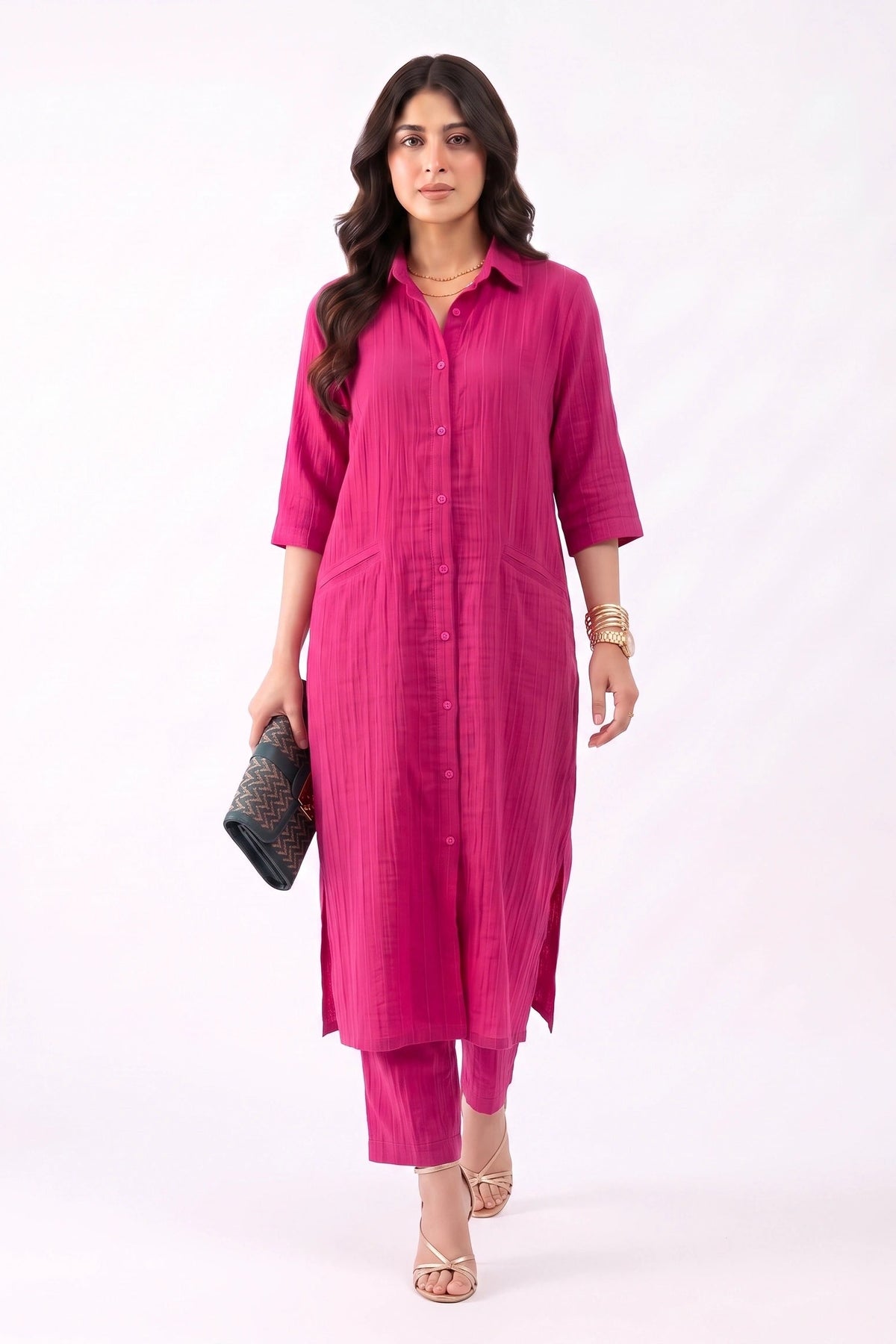 A woman modeling a bright pink three-piece kurta set, featuring a long button-up shirt-style tunic, matching pants, and holding a black clutch bag against a plain white studio background.