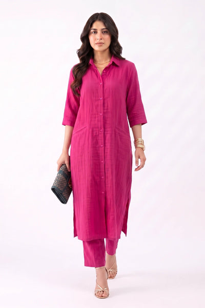 A woman modeling a bright pink three-piece kurta set, featuring a long button-up shirt-style tunic, matching pants, and holding a black clutch bag against a plain white studio background.