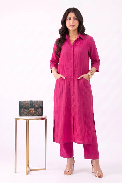 A woman models a bright pink long kurta and matching pants set, standing next to a small gold-framed table holding a black patterned clutch bag, against a plain white studio background.