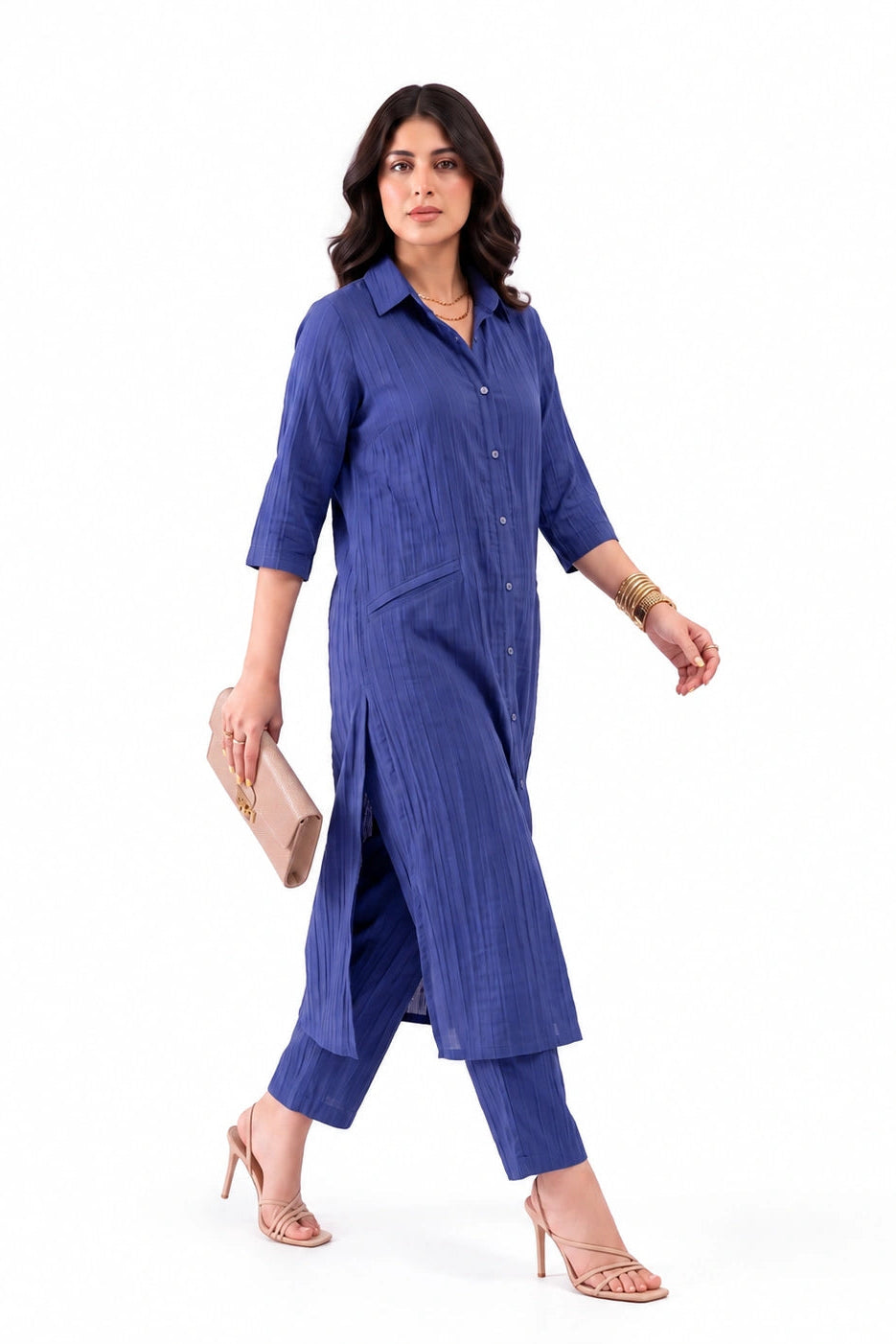 A woman modeling a blue button-front tunic with matching cropped pants, shown against a plain white studio background. She is holding a beige clutch and wearing beige high-heel sandals.