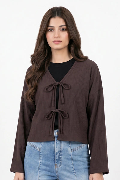A woman modeling a dark brown long-sleeve cropped top with a V-neck and tie-front detail, paired with light blue jeans and holding a small white handbag, against a plain white studio background.