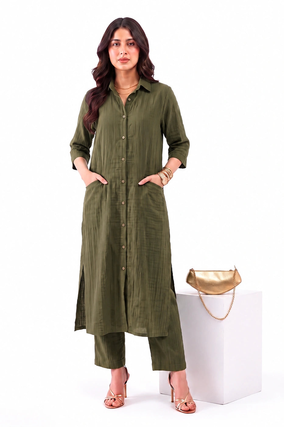 A woman models an olive green long kurta with matching pants, standing next to a white block displaying a gold clutch bag against a plain white background.