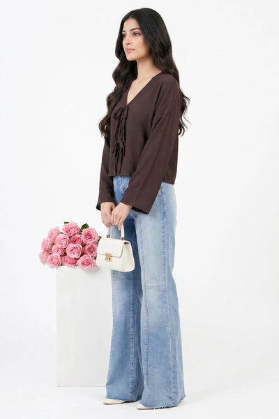 A woman stands in a studio wearing a dark brown long-sleeve tie-front blouse and light blue wide-leg jeans, holding a small white handbag next to a white pedestal with pink flowers.