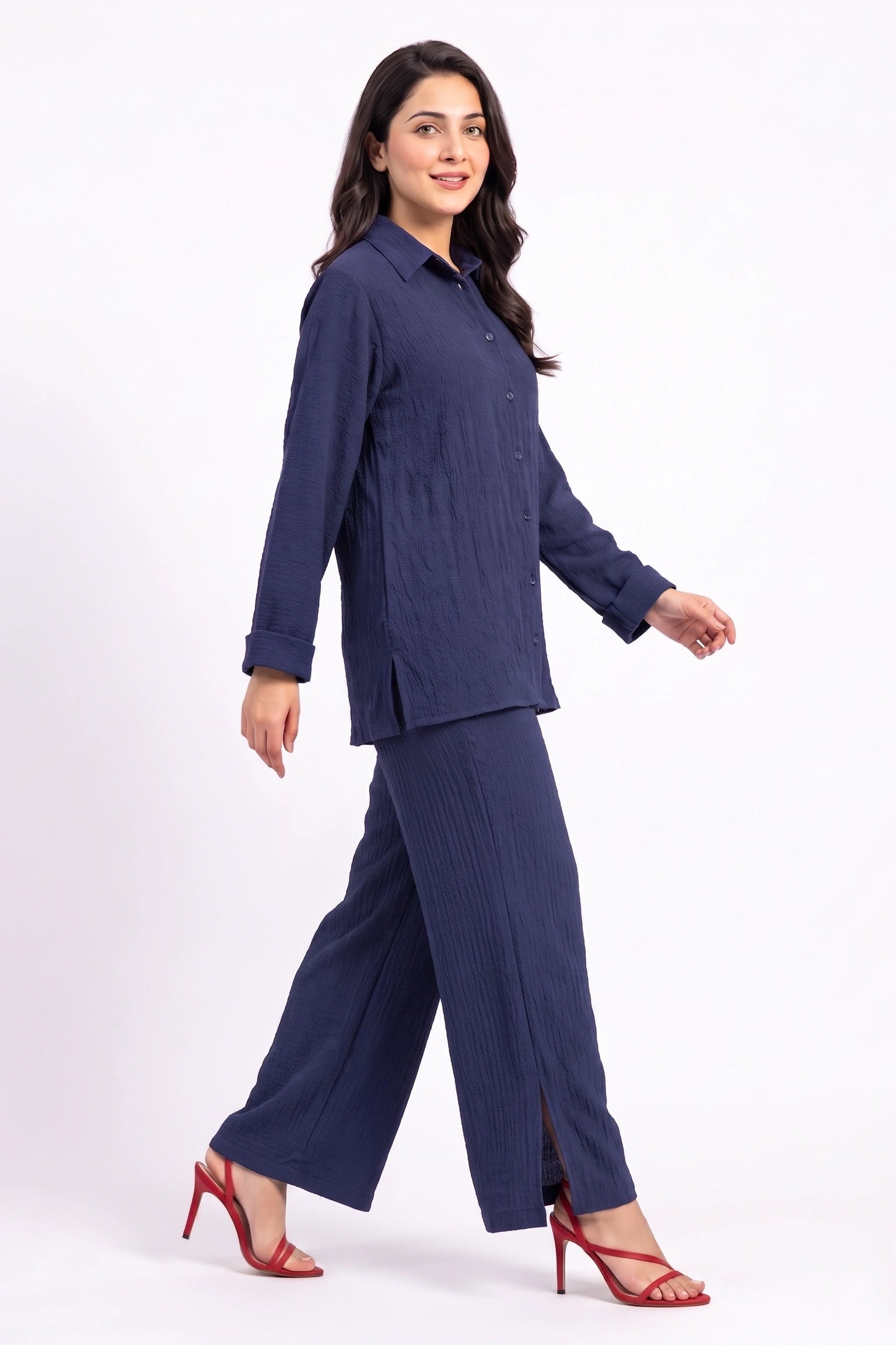 A woman modeling a navy blue two-piece set consisting of a long-sleeve button-up shirt and matching wide-leg trousers, paired with red high-heeled sandals, against a plain white studio background.