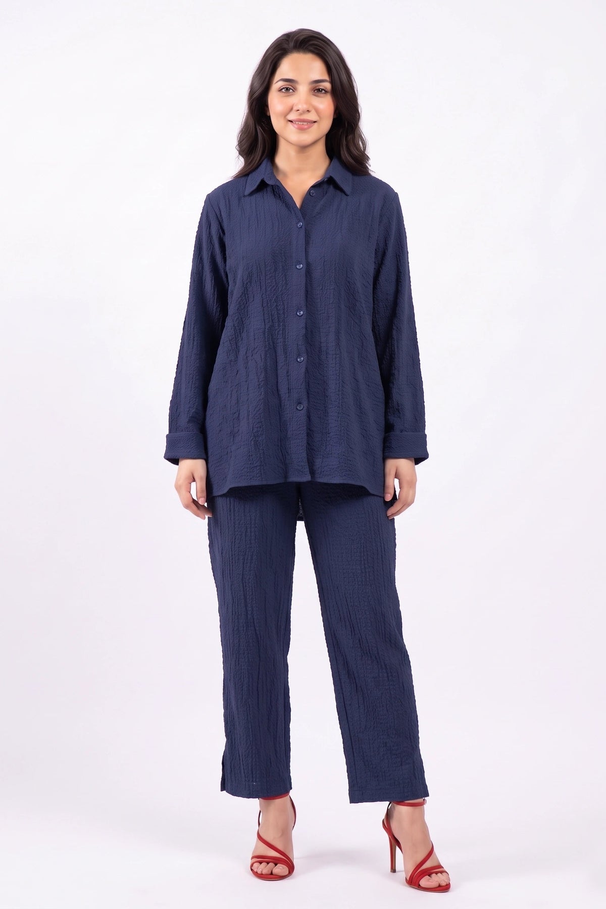 A woman modeling a matching navy blue two-piece set consisting of a long-sleeve button-up shirt and wide-leg cropped pants, styled with red high-heeled sandals against a plain white studio background.