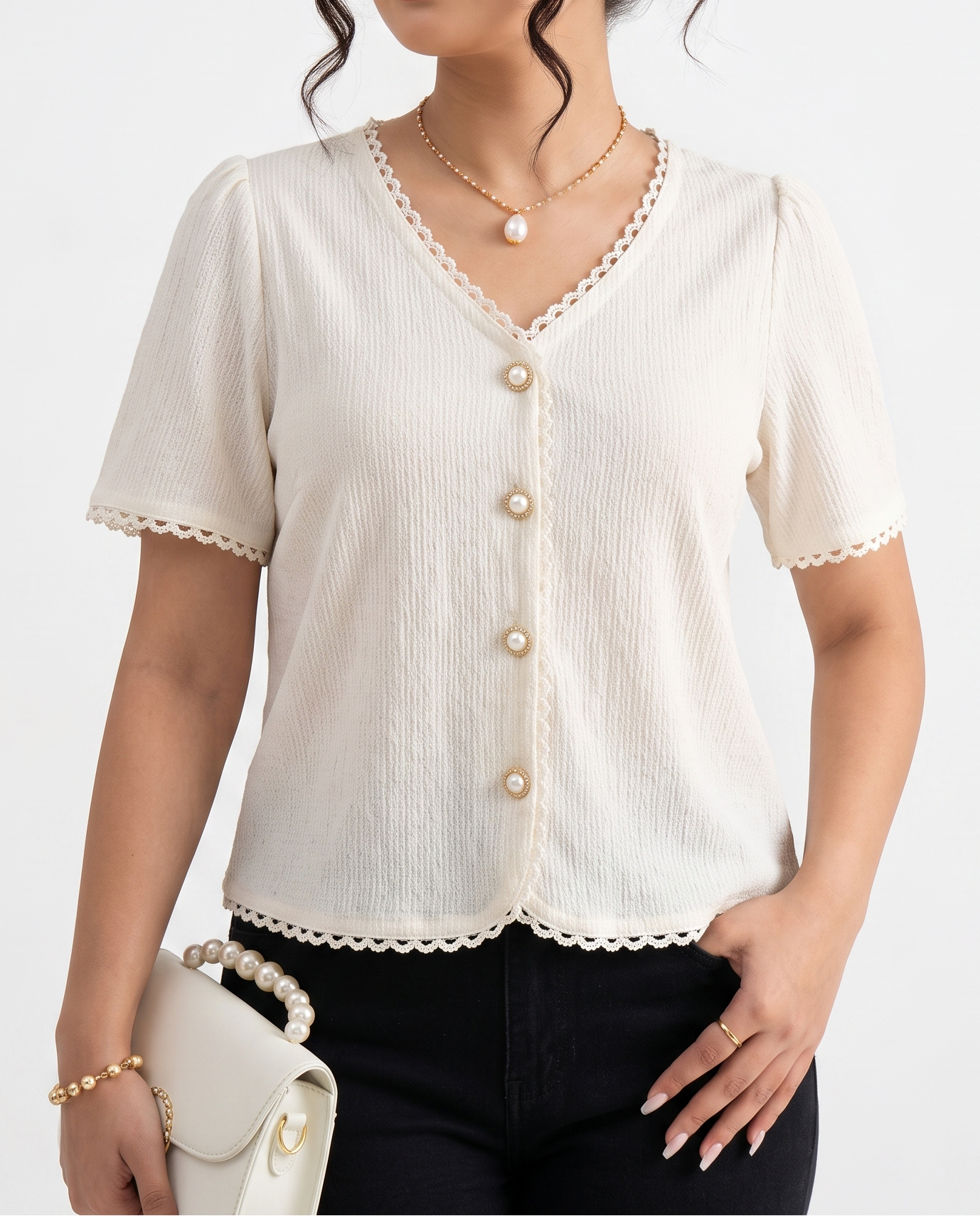 A woman modeling a short-sleeve cream-colored top with scalloped edges and a button-up front, paired with black pants and holding a small cream handbag against a plain white background.
