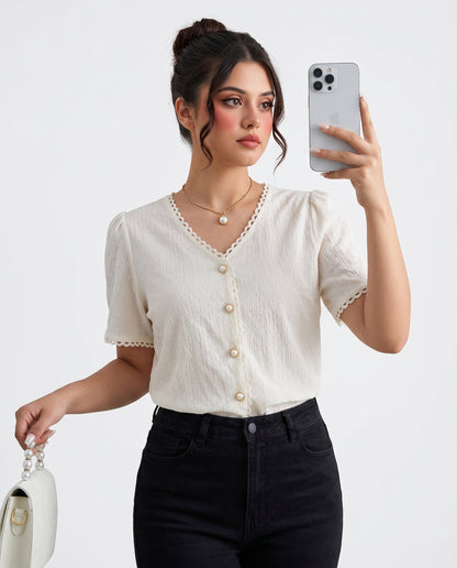 A woman takes a mirror selfie wearing a short-sleeve cream-colored textured top with scalloped edges and decorative buttons, paired with black jeans.