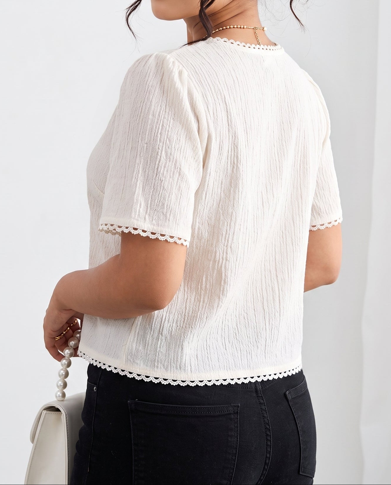A woman wearing a short-sleeve textured cream top with delicate lace trim on the sleeves and hem, paired with black jeans, shown from a three-quarter back view against a white curtain background.