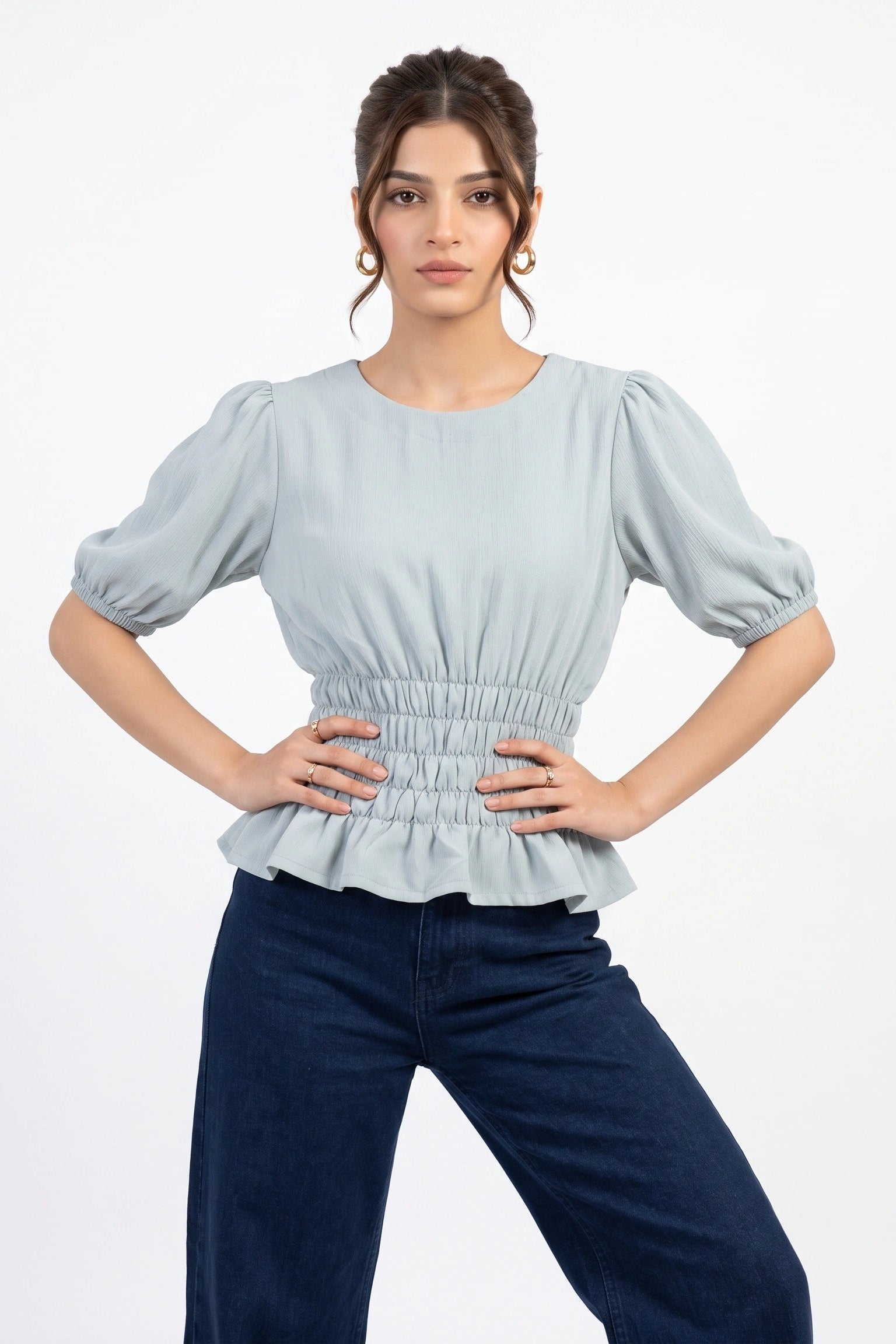 A woman modeling a light blue puff-sleeve top with a smocked waist and peplum hem, paired with dark blue jeans, against a plain white studio background.