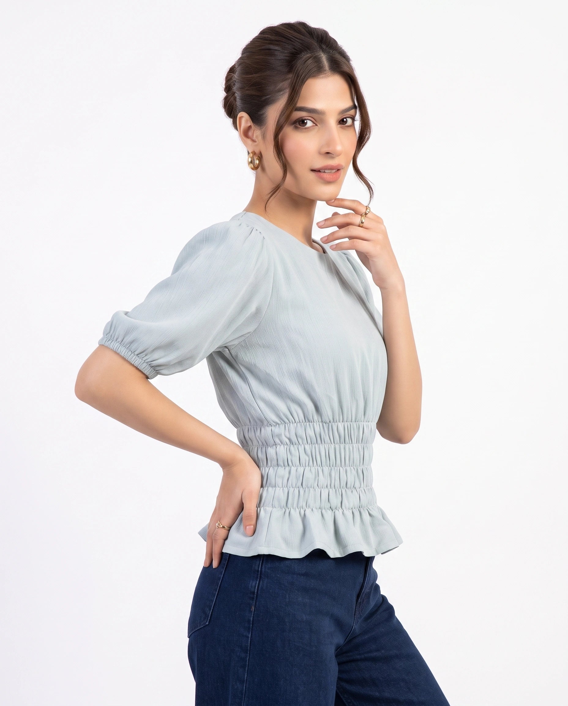 A woman modeling a light blue-gray short-sleeve blouse with puff sleeves, a smocked waist, and a ruffled hem, paired with dark blue jeans against a plain white background.