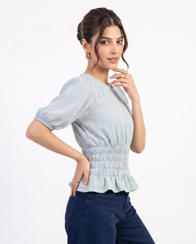 A woman modeling a light blue-gray short-sleeve blouse with puff sleeves, a smocked waist, and a ruffled hem, paired with dark blue jeans against a plain white background.