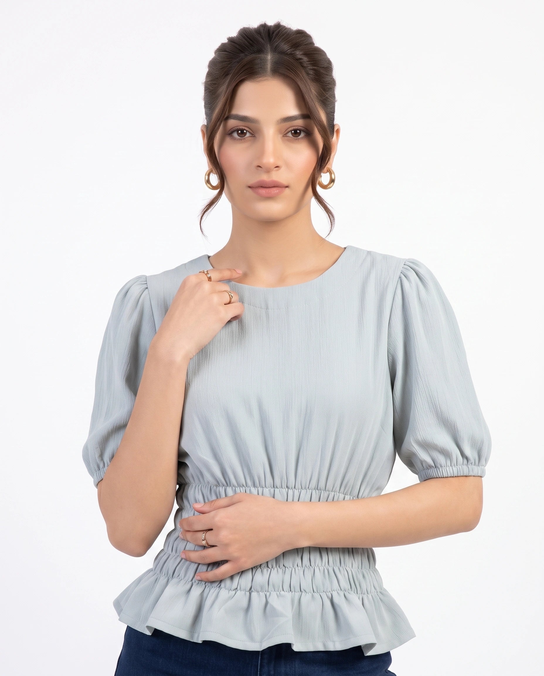 A woman modeling a light blue-gray puff-sleeve top with a gathered elastic waist and peplum hem, paired with dark blue jeans, against a plain white background.