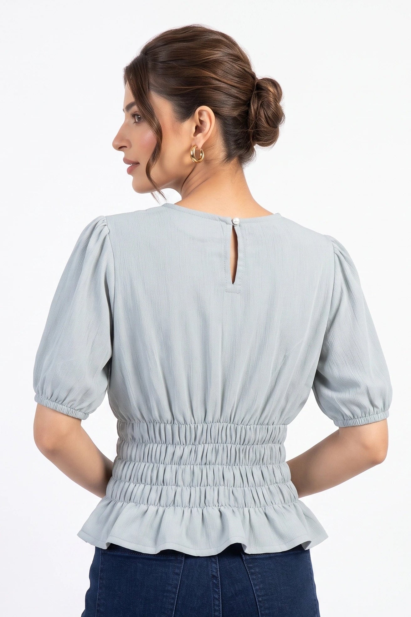 A woman shown from the back wearing a light blue-gray short-sleeve blouse with puff sleeves, a smocked waist, and a ruffled hem, paired with dark blue jeans against a plain white background.