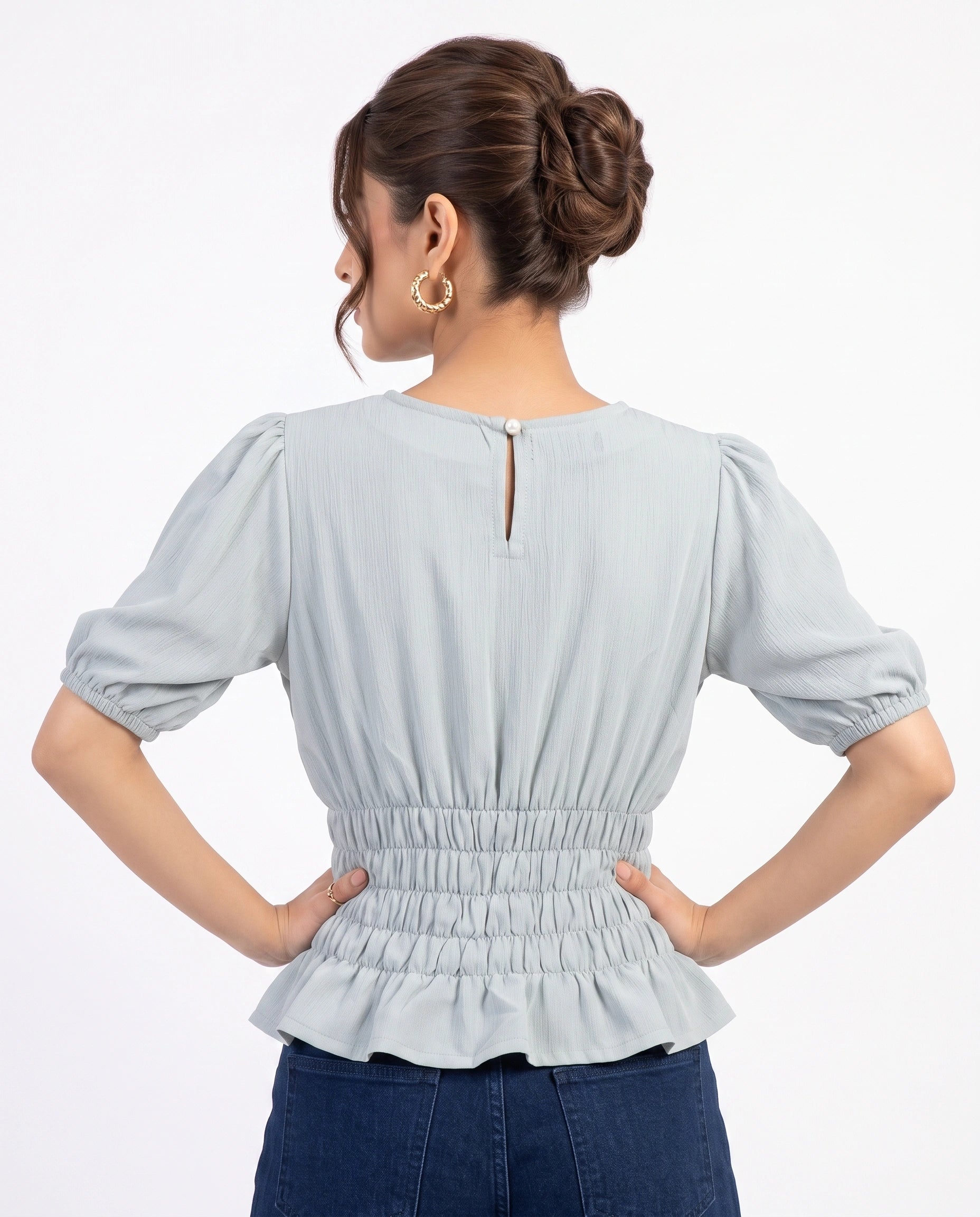 A woman shown from the back wearing a light blue-gray short-sleeve blouse with puff sleeves, a gathered waist, and a peplum hem, paired with dark blue jeans against a plain white studio background.