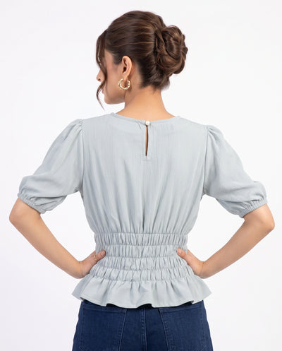 A woman shown from the back wearing a light blue-gray short-sleeve blouse with puff sleeves, a gathered waist, and a peplum hem, paired with dark blue jeans against a plain white studio background.