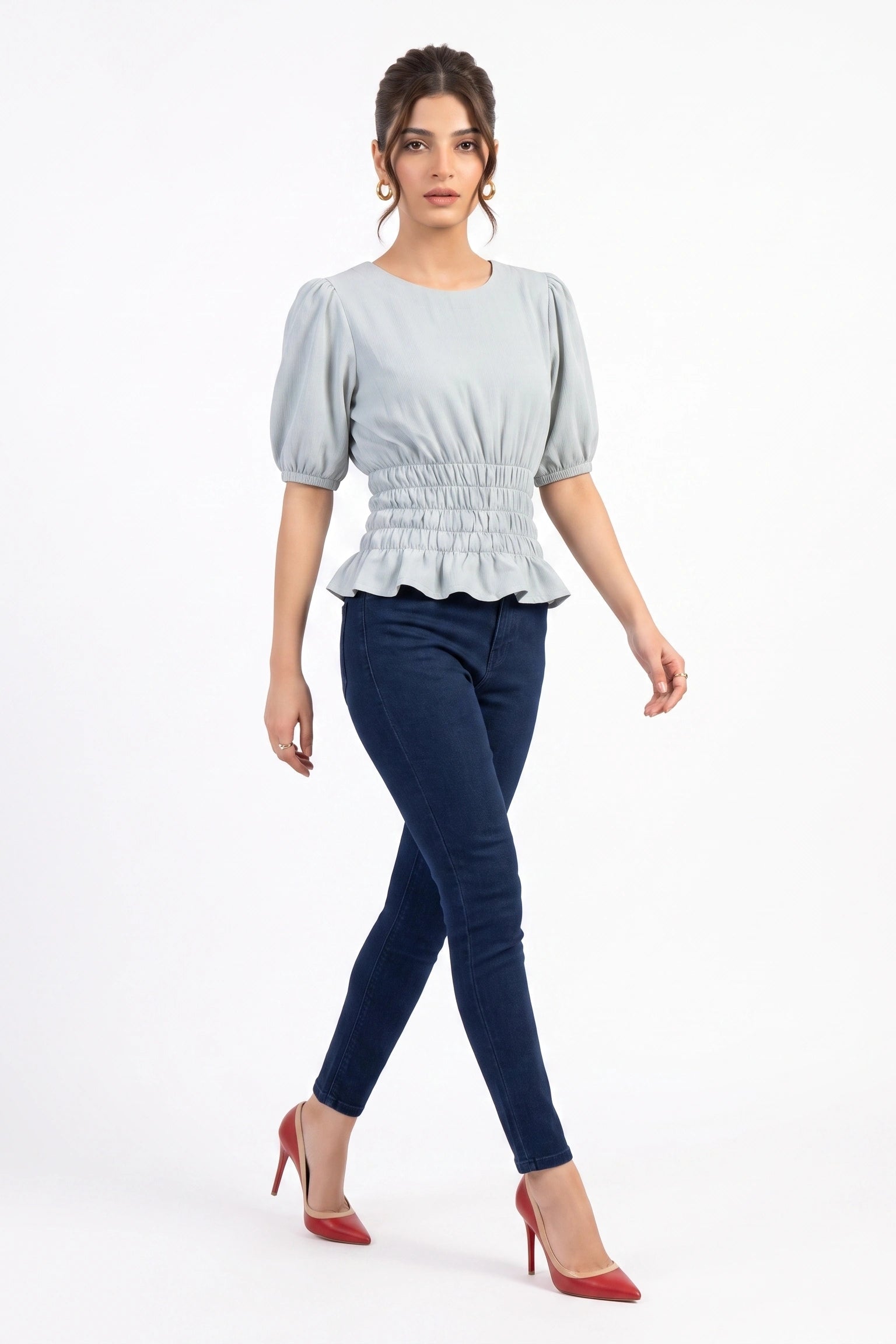 A woman modeling a light blue-gray short-sleeve blouse with puffed sleeves and a smocked waist, paired with dark blue skinny jeans and red high heels, against a plain white studio background.