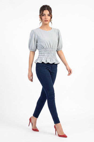 A woman modeling a light blue-gray short-sleeve blouse with puffed sleeves and a smocked waist, paired with dark blue skinny jeans and red high heels, against a plain white studio background.
